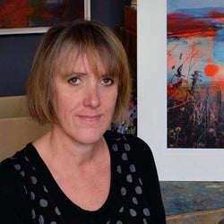 An image of Kate, a white woman with short light brown hair. She is wearing a black top with grey polka dots. In the background is some of her artwork.