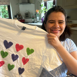 An image of Sophia, who has dark hair and is wearing a light blue top. She is smiling and holding up one of her t-shirts, which is white with multi-coloured hearts.