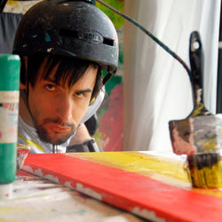 An image of Michael, a white man with dark brown hair and a short beard, leant over a canvas. He is wear a black helmet which has a paint brush attached which he is using to paint.