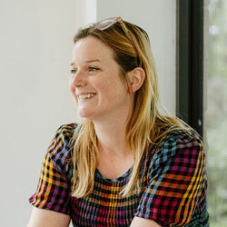 An image of Badbidu. She is facing away from the camera smiling. She is a white woman with long blonde hair, wearing a multi-colour check top with glasses perched on her head.