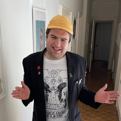 An image of Johnny smiling with his hands open wide. Johnny is a white man with short light brown hair. He is wearing a black blazer with pin badges, a graphic t-shirt, and a yellow cap.